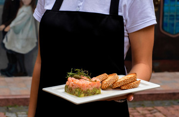  a girl in a black apron and a white bozka is holding in front of her a salmon avocado and rucola appetizer, .white bread cracker on a white plate. background street