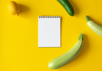 Empty white paper notepad, zucchini, lemon and cucumber on yellow background with space for text....