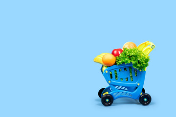 A grocery shopping cart full of salad, oranges, bananas, apples and bread. Close-up concept with place for text on a blue background.