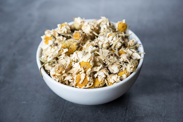 Dried chamomile tea in white porcelain bowl on stone, concrete background.  Dry, organic, healthy and relaxing herbals.