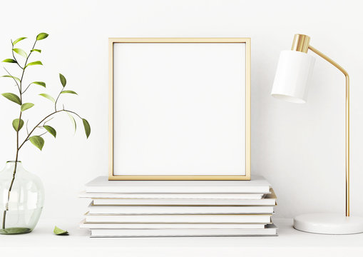 Interior poster mockup with square gold metal frame on pile of books, green tree branch in vase and desk lamp on empty white wall background.  3D rendering, illustration.