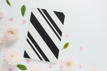 .black and white striped notebook, pink plastic hearts and pink flowers of chrysanthemums and green leaves on a white table. space for text..
