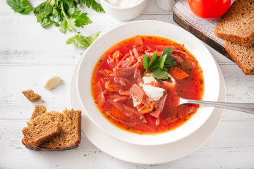 white plate of Ukrainian red borscht with parsley and sour cream, a spoon, pieces of bread on a white wooden background