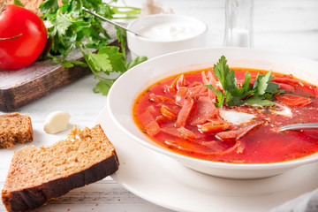 plate of Ukrainian red borscht with parsley and sour cream, a spoon, pieces of bread on a white wooden background, closeup