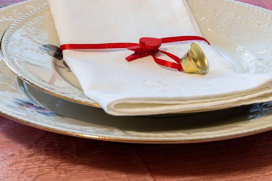 Preparations for the end of year dinner at home. A set table with a napkin closed by a small heart-shaped clothespin and a golden bell on two porcelain plates.