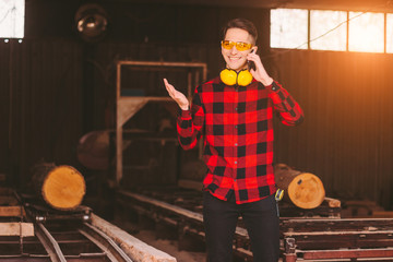 Young happy sawmill workman in protective glasses and headphones talking cell phone while working...
