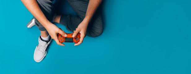 A young guy plays video games in his hands holding a red gamepad on a blue background, sitting in gray sneakers, long banner © Anastassiya 