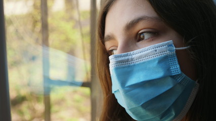 Close-up of a girl in a protective medical mask on her face sadly looks out the window while staying at home in quarantine - obrazy, fototapety, plakaty