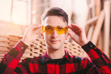 Happy man cabinet maker wearing protective glasses and smiling while working at sawmill factory. Young male carpenter hold eyewear protection at woodworking workshop. Professional personal protection