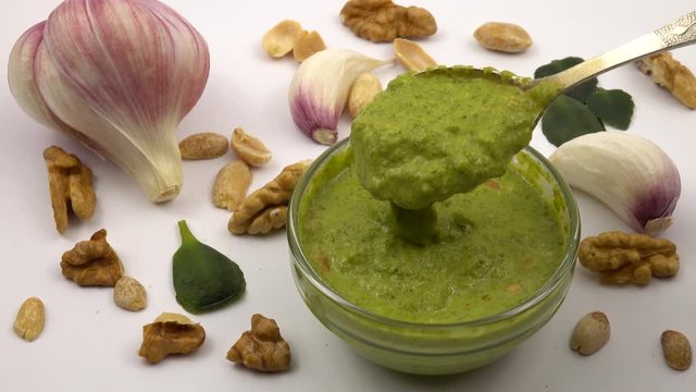 Pesto sauce flows from a spoon into a small glass bowl. Glass bowl with pesto sauce on a background of walnut kernels and garlic