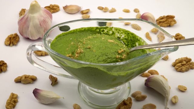 Pesto sauce flows from a spoon into a gravy boat. Gravy boat with pesto sauce on a background of walnut kernels and garlic