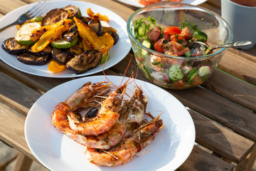 grilled langoustines and vegetables on white plates, a bowl with fresh salad on a garden wooden table outdoor. Summer time