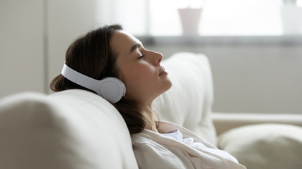 Peaceful girl in modern wireless headphones sit relax on comfortable couch listening to music,...