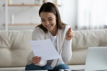 Excited young woman sit on couch feel satisfied joyful reading good news in paper bank letter...