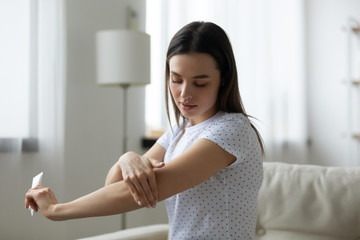 Young Caucasian woman sit on couch in living room using moisturizing body cream for soft healthy...