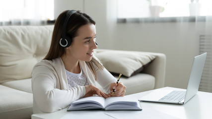 Happy millennial girl in headphones make notes watch webinar on laptop, smiling young woman in headset taking online course or web training, study on computer at home, distant education concept