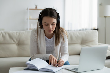 Concentrated millennial girl in headphones sit on couch at home make notes studying online on laptop, focused young woman watch webinar or Internet course on computer, distant education concept