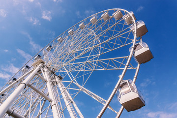 White metal construction of an attraction Ferris wheel with observation cabs high against a blue sky with white clouds.