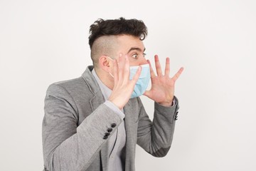 Portrait of young man wearing medical mask with shocked facial expression, holding hands near face, screaming and looking sideways at something amazing.