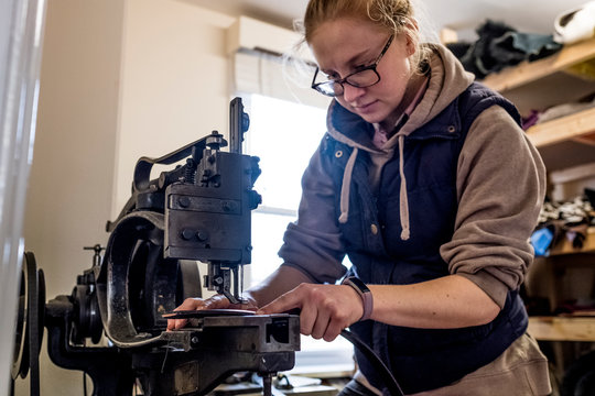 Female saddler standing in workshop, sewing leather strap on saddlery sewing machine.