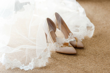 Beige bridal shoes standing on the floor, covered with a white veil with lace. A pair of expensive and beautiful wedding shoes with high heels, with gold and silver finish