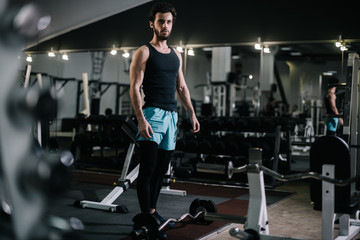 Fototapeta na wymiar Athletic bearded young man with muscular wiry body wearing sportswear posing in modern dark gym. Concept of healthy lifestyle.