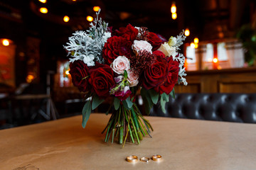 Three gold wedding rings with gemstone and bridal bouquet of red flowers lying on the table in the restaurant in loft style