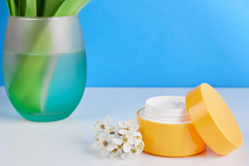 Jar of cosmetic cream and soft green vase with flowers on pink background. Natural cosmetic product with herbs.
