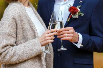 Bride in white dress and coat and groom in blue suit hold in their hands wineglasses with champagne and hugging. Wedding bounquet and celebration