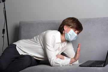 Obraz premium Young woman in medical protective mask lying on a grey couch and waving with her hand, looking on a computer display. Home activity, isolation and pandemic concept