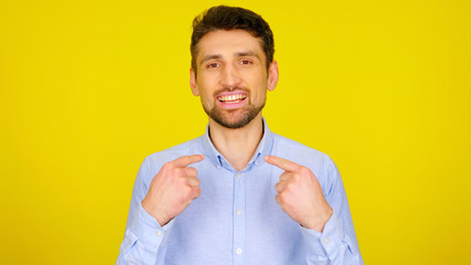 Choose me! Handsome bearded man shows his fingers on a yellow background with copyspace. Guy in a light blue shirt with his hands up. Place for text or product - obrazy, fototapety, plakaty