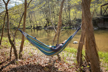 Young man is lying in a colorful hammock  near the trees, a river and mountainse early in the spring. He is alone and have a relaxing time in the nature