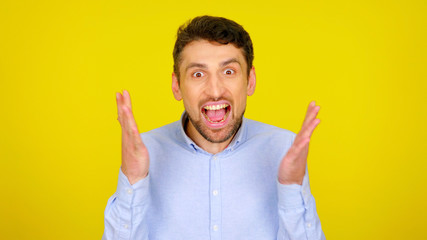 Emotional man with open mouth in a light blue shirt on a yellow background with copyspace. Handsome bearded guy screams and rejoices in victory for his beloved team. Place for text or product