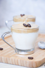 Close up of a crystal glass with milk and whipped instant coffee with raw coffee beans on top on a bamboo tray and a spoon and a white background with depth of field. Dalgona coffee concept