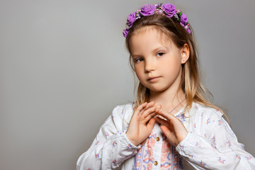 Portrait of posing pretty little girl in white dress and purple wreath on gray isolated background . 