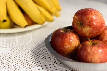 Close-up de Maças dentro de uma vasilha de metal ao fundo, um cacho de bananas sobre um prato.