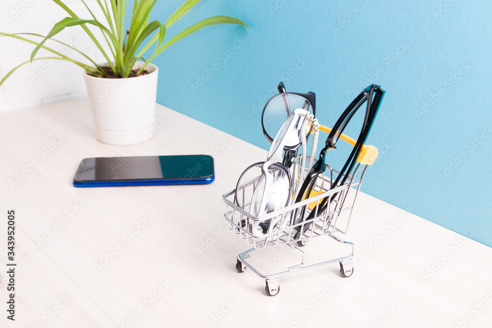 Wall mural children's glasses and two pairs of black glasses of adults in a little shopping trolley on a blue background, online purchase of glasses, a flower and a smartphone in the background in blur