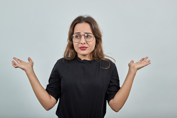 Cute young brunette woman in black t-shirt, blue jeans with belt on gray background, confused girl with glasses shocked by the news, spread her arms wide