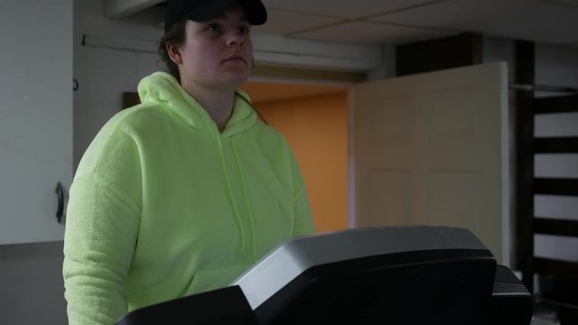 Caucasian Young Woman Is Walking Slowly On Her Treadmill In Basement Of Residential Home - Front Angle 1