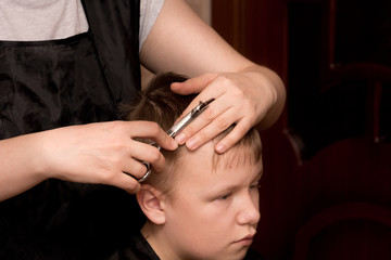 a mother-hairdresser in a black apron with a comb and scissors in her hands at home does her hair for her son, who is sitting on a chair and wrapped in a black blanket. quarantine self-isolation