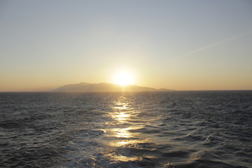 An island in the sea absorbed by the rays of sunset, its outlines are lost in the haze and in the water there are golden reflections.