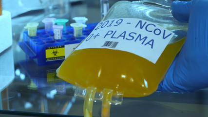 A gloved had of a medical technician holding an IV bag of blood plasma for possible treatment of...