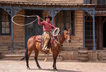 Cowboy using lasso while riding his horse through an old western town - obrazy, fototapety, plakaty