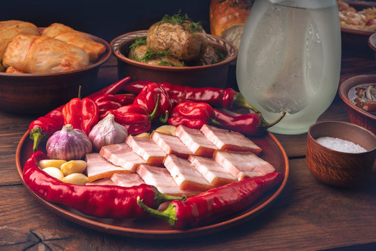 Traditional Ukrainian food - a plate with lard, garlic and hot pepper, pickled cucumbers and tomatoes in a clay bowl, donuts, a bottle of vodka and a glass on a dark wooden background.
