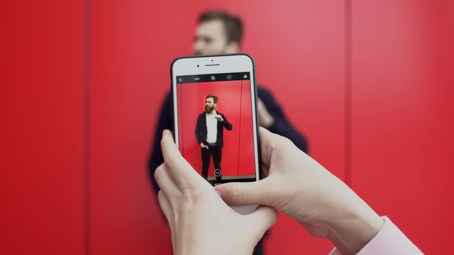 Person is taking pictures of posing near red wall brunette bearded man on the phone. Young funny guy is posing for photographer with smartphone against red background.