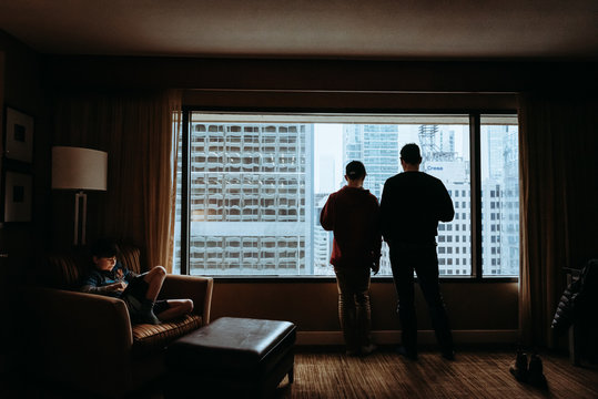 Father and son looking through window while brother sitting on armchair at home