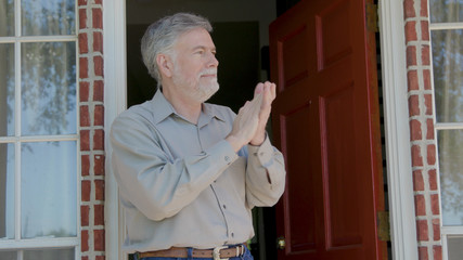 A mature man stands on the front porch of his home during the COVID19 pandemic of 2020 applauding brave healthcare workers who are off scene.