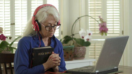 An older woman holding a Bible and praying as she attends church virtually via her laptop due to congregation and group fellowship restrictions during COVID19 pandemic.