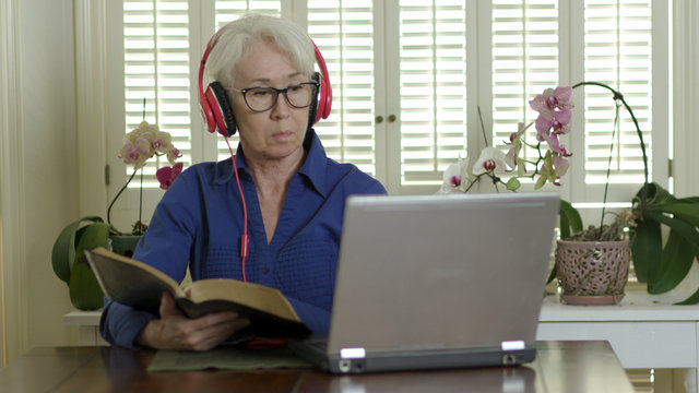 A mature woman studies the Bible as she attends church virtually by streaming it to her laptop due to congregation and group fellowship restrictions during COVID19 pandemic.