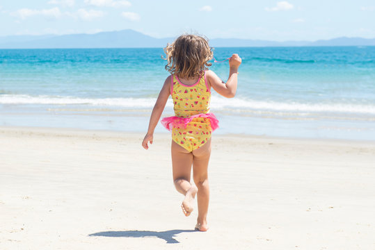 Back view of girl running in swimsuit on the beach towards the blue sea on beautiful sunny day.